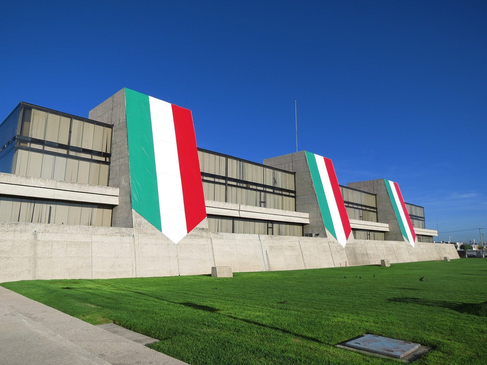 Edificio del INEGI en Aguascalientes con tres banderas de México colgadas en la fachada de concreto, sobre un jardín de césped
